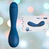 Image showcasing a blue, curved OhMiBod NEX2 BlueMotion G-Spot Vibrator - 2nd Gen with "+" and "♥" buttons near the base. Next to it is a box labeled "OhMiBod NEX2 BlueMotion," highlighting the product's features such as app-controlled bliss and depicting the same massager on the packaging. The background features a soft-focus light bokeh.